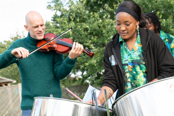 3 musicians playing outside. Two playing the steel dreams, the other a viola. A large green tree is in the background