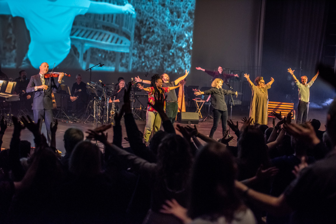 A stage full of performers with their arms raised an smiling. An audience looks on with their arms also raised