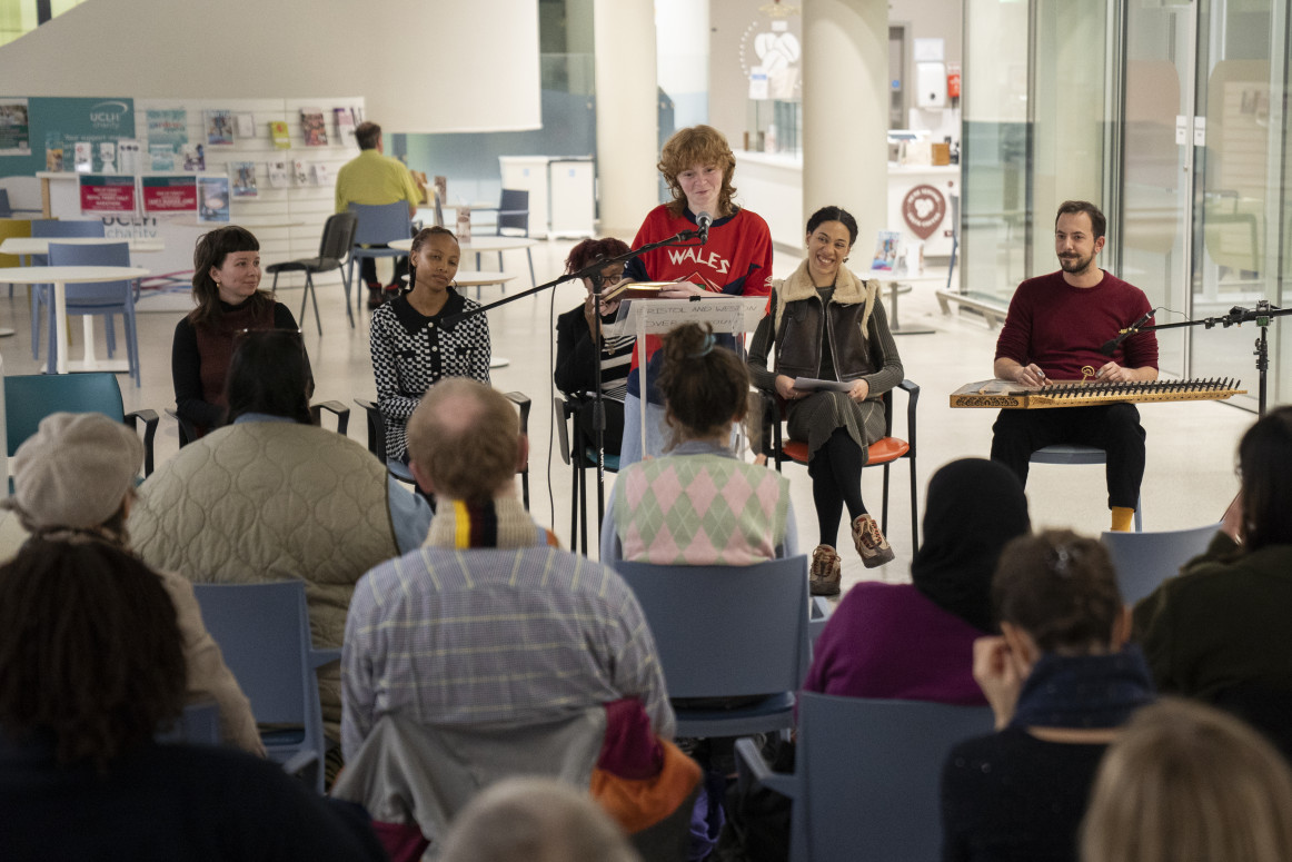 A poet standing performs in front of a seated audience with their backs to the camera. Behind the poet four other performers are seated smiling and looking on