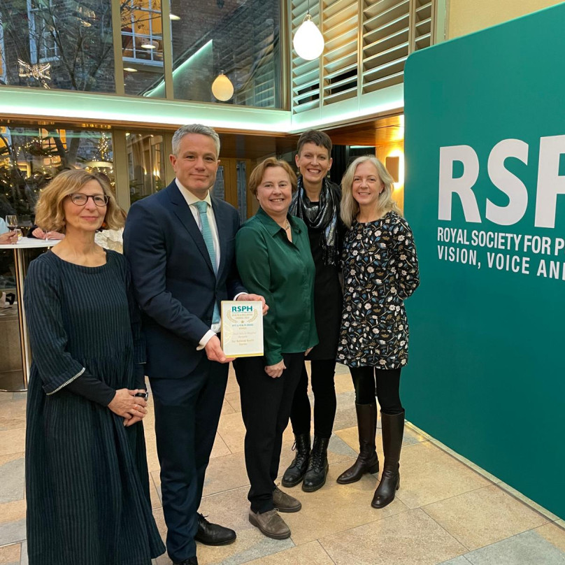 A group of 4 people smiling, one is holding an award. They are standing next to a large green banner that says RSPH Vision, Voice and Practice